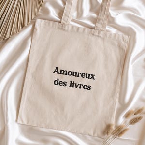 Op de afbeelding: Een beige canvas draagtas met de zwarte tekst "Amoureux des livres". De tas heeft twee handvatten en ligt op witte satijn. Gedroogde planten en een decoratieve waaier zijn ook zichtbaar.
