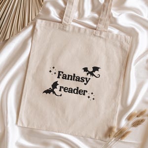 Peut inclure: Un sac fourre-tout en toile beige avec les mots "Fantasy reader" et deux silhouettes de dragons noirs. Le sac a deux poignées beiges et est posé sur un fond de satin blanc.