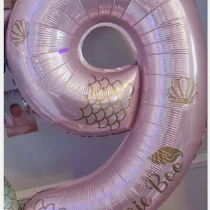 May include: A large, pink number nine balloon with gold seashell and scale designs. The balloon has the name "Bessie Boo" printed on it. Several gold and pink balloons are also present, creating a festive birthday theme.
