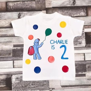 May include: White t-shirt with a cartoon character holding a green balloon. The text "Charlie is 2" is printed in blue. The shirt is decorated with colorful polka dots. The character is blue and holding a red bag.