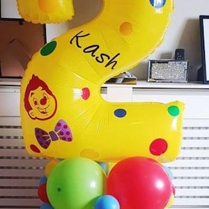 May include: A large yellow number 2 balloon, decorated with colourful spots and the name "Kash". It features a cartoon character and sits atop a base of mixed balloons. The base includes red, green, and yellow balloons. Party decoration.