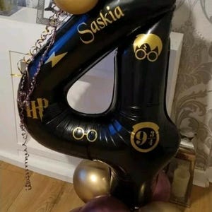 May include: A black number 4 balloon with gold detailing, personalised with the name "Saskia" and Harry Potter symbols. The balloon is part of a display with gold and purple balloons, ideal for a birthday celebration.