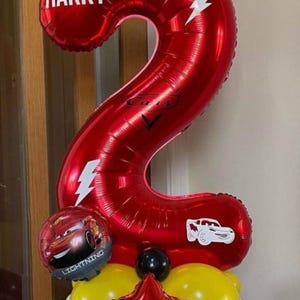 May include: A large red number 2 balloon with the name "HARRY" in white, featuring lightning bolt and car motifs. The balloon display includes a "Lightning McQueen" balloon and a cluster of yellow and red balloons.