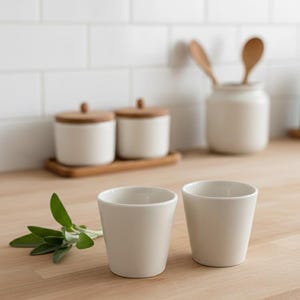 May include: Two off-white ceramic cups sit on a light wood surface, with a sprig of green leaves to the left. In the background are white ceramic containers with wooden lids and a jar with wooden spoons, suggesting a kitchen or dining theme.