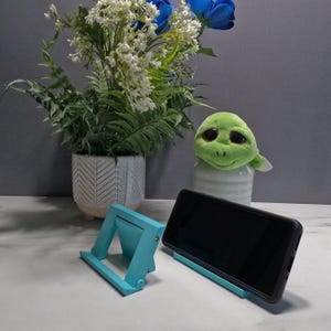 May include: A teal-colored phone stand with a triangular design, holding a black smartphone. Another teal stand is visible. A plush green turtle and a potted plant with white and blue flowers are in the background.