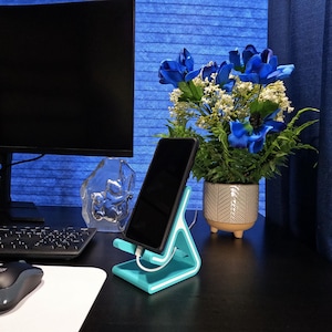 May include: A teal and white phone stand with a smartphone charging. The stand has a modern, geometric design. A black computer monitor, keyboard, and mouse are visible in the background. A vase of blue flowers is also present.