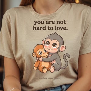 May include: Beige t-shirt with a cartoon illustration of two hugging monkeys. The text "you are not hard to love" is printed above the monkeys. The phrase "emotional support energy" is printed below the monkeys.