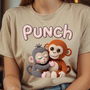 May include: A beige t-shirt featuring the word "PUNCH" in pink embroidered letters. Below the text are two crocheted monkeys, one grey and one orange, embracing. The monkeys have large eyes and are smiling.