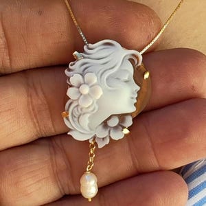 May include: A gold necklace featuring a cameo pendant. The pendant showcases a white carved profile of a woman with flowers, set against a brown background. A small pearl dangles from the bottom of the pendant.