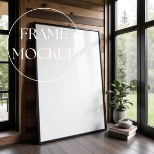 Op de afbeelding: Een grote mockup van een kunstafdruk met zwarte lijst, leunend tegen een houten wand. De tekst "FRAME MOCKUP" staat over de witte afdruk. Een kleine plant in een pot en boeken staan op de houten vloer naast een groot raam.