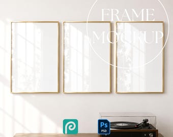 May include: Three gold-framed rectangular mockups with white interiors hang on a white wall. The text "FRAME MOCKUP" is overlaid on the frames. A wooden cabinet with a record player and design software icons are below the frames.