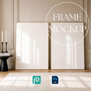 May include: Two large vertical rectangular frames with thin gold borders standing side by side. The frames contain blank white surfaces. To the left, a black pedestal table holds two white candles and stacked books. Text reads FRAME MOCKUP.