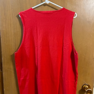 May include: A sleeveless, bright red tank top hanging on a white hanger. The top has a scoop neck and is made of a soft, solid-colored fabric. The garment is displayed against a light brown wooden door.