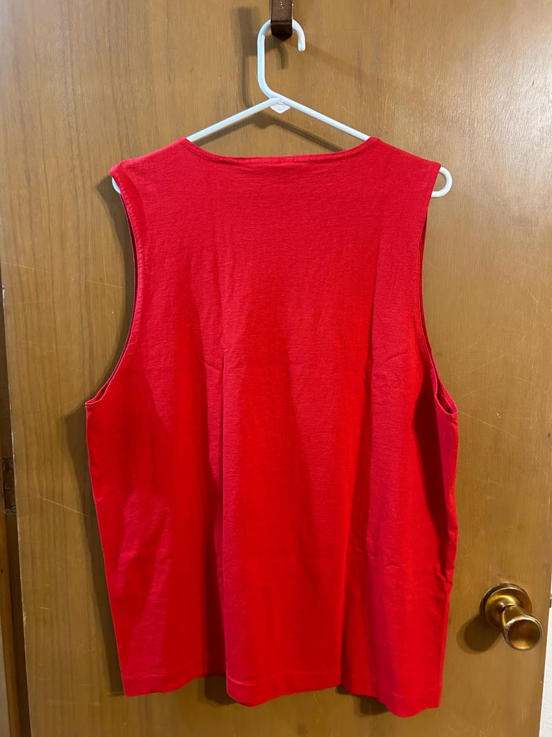 May include: A sleeveless, bright red tank top hanging on a white hanger. The top has a scoop neck and is made of a soft, solid-colored fabric. The garment is displayed against a light brown wooden door.