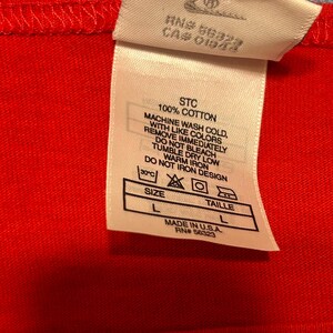 May include: Close-up of a red garment with a white tag. The tag includes washing instructions, size information, and the text "MADE IN U.S.A." The garment is 100% cotton. The size is L.