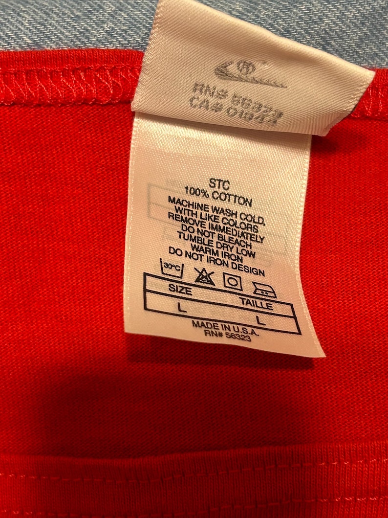 May include: Close-up of a red garment with a white tag. The tag includes washing instructions, size information, and the text "MADE IN U.S.A." The garment is 100% cotton. The size is L.