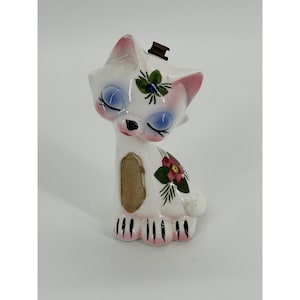 May include: A white ceramic cat figurine with closed eyes, pink-tinted ears, and a small dark metal component on top of its head. The cat features a painted green floral design on its forehead and a red flower on its side, with black paw markings.