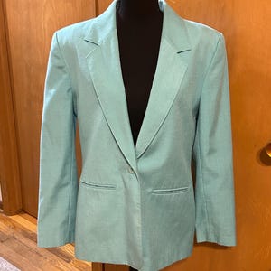 May include: A light turquoise blazer with a single-button closure and two front pockets. The blazer is displayed on a black mannequin. The jacket has a classic lapel design and long sleeves.