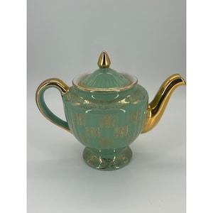 May include: A mint green ceramic teapot featuring a gold metallic handle, spout, and finial. The body of the teapot is decorated with a repeating gold square pattern and vertical ribbing, resting on a pedestal base with gold trim accents.