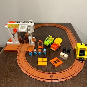 May include: A Fisher-Price Lift & Load Railroad playset. The set includes a white and orange loading station, a brown track, a yellow train car, two small trucks, and several accessories. The text "Lift & Load Railroad" is visible on the loading station.