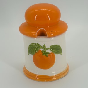 May include: A ceramic jar with an orange lid and a white body. The jar features a painted orange fruit with green leaves. The lid has a small opening. The jar is designed for kitchen use.