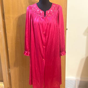 May include: A vibrant fuchsia-colored robe displayed on a black mannequin. The robe features long sleeves with ruffled cuffs and a delicate floral lace detail around the neckline. It is buttoned down the front.