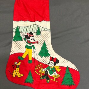 May include: Red Christmas stocking featuring Mickey Mouse pulling Minnie Mouse on a sled, with Pluto nearby. The background has green Christmas trees and a snowy landscape. The top of the stocking is red.