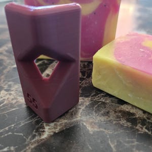 May include: A dark purple, L-shaped plastic corner guide with a diamond-shaped cutout and the number 55 embossed on the front, positioned next to a rectangular block of yellow and pink marbled soap with visible dark speckles throughout the surface.