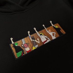 Op de afbeelding: Zwarte hoodie met een geborduurd embleem met stripfiguren met opgeheven armen. De rechthoekige patch heeft een achtergrond van houten planken. De hoodie heeft een capuchon en een label "KEEPNEEDLE Made in Ukraine".
