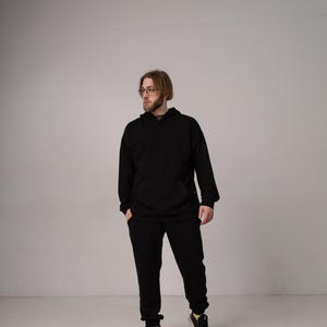 Pu&ograve; includere: Una persona indossa una felpa con cappuccio nera e pantaloni della tuta abbinati. La felpa ha una tasca frontale e un cappuccio con coulisse. I pantaloni della tuta hanno una vestibilit&agrave; ampia. La persona indossa anche scarpe da ginnastica nere con dettagli rosa e gialli.