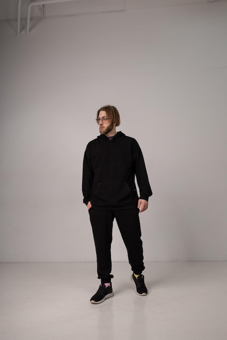 Pu&ograve; includere: Una persona indossa una felpa con cappuccio nera e pantaloni della tuta abbinati. La felpa ha una tasca frontale e un cappuccio con coulisse. I pantaloni della tuta hanno una vestibilit&agrave; ampia. La persona indossa anche scarpe da ginnastica nere con dettagli rosa e gialli.