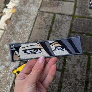 Może przedstawiać: Haftowana naszywka przedstawiająca oczy w stylu anime z szarymi i czarnymi włosami. Prostokątna naszywka ma szarą obwódkę. Oczy są szczegółowe z białymi refleksami i zdecydowanym wyrazem. Naszywka jest trzymana na tle szarych kostek brukowych.