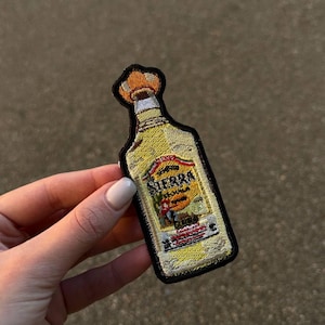 May include: Embroidered patch featuring a tequila bottle with the text "Sierra Tequila". The patch has a yellow and orange color scheme with a black border. The patch is a fun accessory for jackets, bags, or other fabric items.