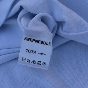 Pu&ograve; includere: Primo piano di un'etichetta bianca di una maglietta con la scritta "KEEPNEEDLE" in nero. Sotto, si legge "100% cotone" con simboli di cura. La maglietta &egrave; realizzata in un tessuto azzurro chiaro.