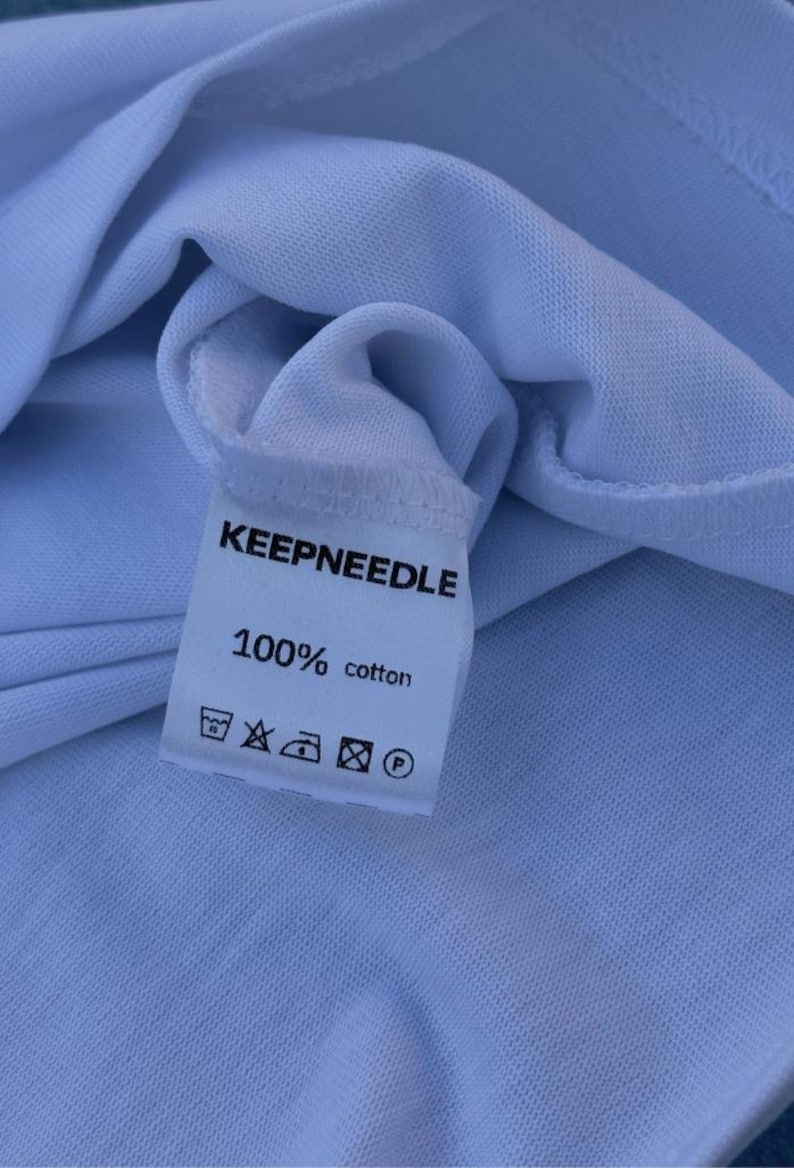 Pu&ograve; includere: Primo piano di un'etichetta bianca di una maglietta con la scritta "KEEPNEEDLE" in nero. Sotto, si legge "100% cotone" con simboli di cura. La maglietta &egrave; realizzata in un tessuto azzurro chiaro.