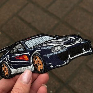 Könnte beinhalten: Gestickter Aufnäher eines marineblauen Sportwagens mit orangefarbenen Rädern und Flammen. Der Aufnäher hat weiße und schwarze Details und ist wahrscheinlich zum Aufbügeln oder Aufnähen auf Stoff gedacht.