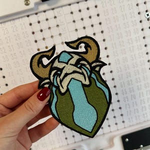 Puede incluir: Parche bordado con la imagen de una cabeza de carnero estilizada con cuernos curvos. El parche presenta una combinación de colores beige, azul y verde. El diseño está detallado con varias texturas y costuras, adecuado para ropa o accesorios.