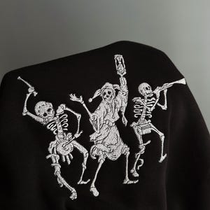 May include: A black sweatshirt featuring a white embroidered graphic of three skeletons. The center skeleton wears a jester hat and holds an hourglass, while the skeleton on the left plays a drum and the one on the right plays a long trumpet.