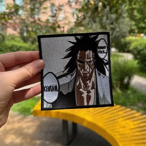 Peut inclure: Un écusson brodé carré présentant une illustration de style manga en noir et blanc d'un personnage aux cheveux noirs hérissés et au visage balafré. L'écusson comprend deux bulles de texte, l'une indiquant "KENPACHI" et l'autre du texte illisible.