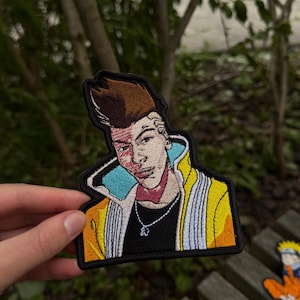 May include: An embroidered patch featuring a character with brown hair, a red mark on the face, and a yellow jacket with white accents. The character wears a black shirt and a silver chain necklace with a small pendant against a black border.