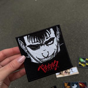 May include: Black square patch featuring a detailed, stylized illustration of a character's face with a sinister grin, rendered in white against a black background. The word "BERSERK" is written in red Japanese characters.