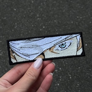 Puede incluir: Parche bordado con un ojo de estilo anime con iris azul claro y cabello blanco. El parche es rectangular con un borde negro. El diseño está sobre un fondo blanco.