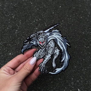 Op de afbeelding: Geborduurde patch met een gedetailleerde illustratie van een krijger in een dynamische pose, die een groot zwaard hanteert. De patch toont een kleurenpalet van grijs, zwart en rood, met ingewikkelde stiksels en een getextureerd uiterlijk. De krijger heeft rode ogen.