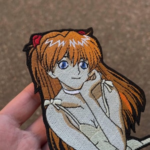 Könnte beinhalten: Gestickter Aufnäher mit einem Anime-Charakter mit orangefarbenem Haar, blauen Augen und einem roten Haaraccessoire. Der Charakter trägt ein helles Outfit mit weißen Schleifen. Der Aufnäher hat einen schwarzen Rand.
