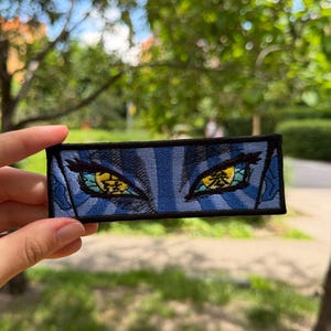May include: A rectangular embroidered patch featuring a pair of stylized blue eyes with dark lashes and yellow irises. The right iris contains a black Japanese kanji character. The design is set against a blue background with a black border.