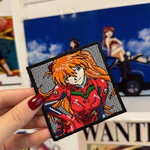 May include: Square embroidered patch featuring a character with orange hair and a red outfit. The patch has a black border and is held by a hand with red nail polish. The background includes posters and artwork.