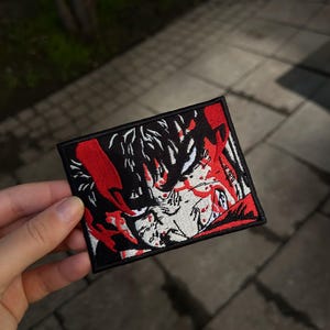 May include: A rectangular embroidered patch featuring a stylized anime character face in black, white, and red thread. The design includes bold black outlines, white facial features, and vibrant red accents, all enclosed within a solid black border.
