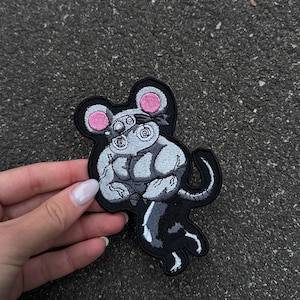 May include: Embroidered patch featuring a stylized gray mouse with a black outline. The mouse has pink ears and a headband. The patch is on a black background and is held by a hand with a light-colored nail polish.