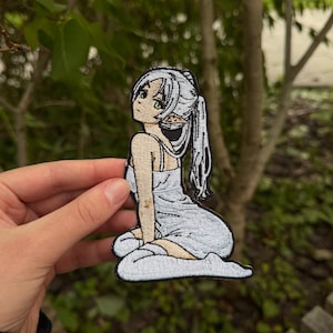 May include: An embroidered patch featuring an anime-style character with long white hair in a ponytail, pointed ears, and a light blue dress. The character is depicted in a kneeling pose, rendered with black outlines and detailed stitching on a fabric base.