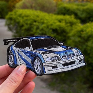 May include: An embroidered patch featuring a white and blue sports car with a black rear spoiler, black windows, and detailed silver wheels. The design includes a prominent front grille, headlights, and blue racing stripes across the hood and sides.